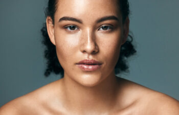 A person with curly dark hair, clear skin, and freckles looks directly at the camera against a plain gray background.