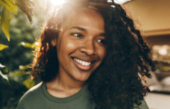 A person with curly hair smiles outdoors in natural sunlight, wearing a green shirt with greenery and sunlight visible in the background.