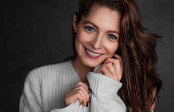 Woman with long brown hair wearing a white sweater, smiling and looking at the camera against a dark background.