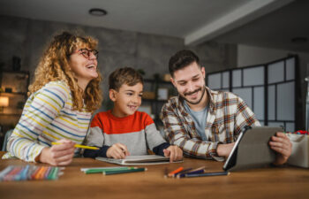 A woman, a boy, and a man sit at a table with colored pencils and a tablet, smiling and working together in a brightly lit room.