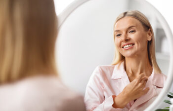 A woman in pink pajamas smiles at her reflection in a round mirror, touching her neck with one hand.