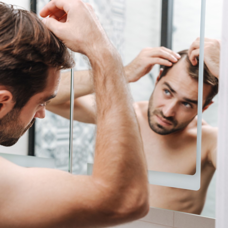 Worried young shirtless man examining his hair while looking at the bathroom mirror