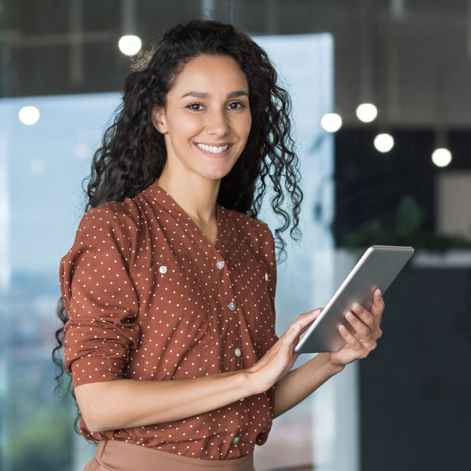 Woman with long curly hair wearing a brown polka dot blouse stands indoors, smiling and holding a tablet.