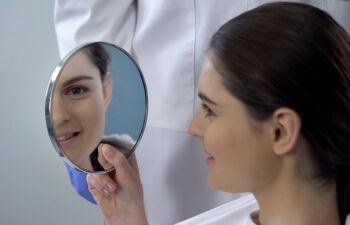 Young female satisfied with rhinoplasty result, smiling face reflected in mirror