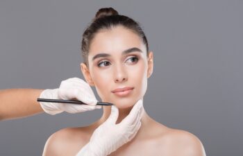 Preparing for plastic surgery. Doctor drawing lines on female face, gray studio background