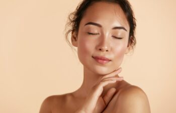 Beauty face. Woman with natural makeup and healthy skin portrait. Beautiful asian girl model touching fresh glowing hydrated facial skin on beige background closeup. Skin care concept
