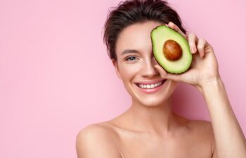 Pretty woman holds half an avocado in front of her face. Photo of attractive woman with perfect makeup on pink background. Beauty & Skin care concept