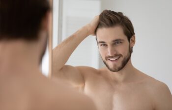 Happy shirtless confident young man looking in mirror combing hair with fingers after morning shower, getting ready for workday or weekend date, feeling satisfied with personal appearance in bathroom.