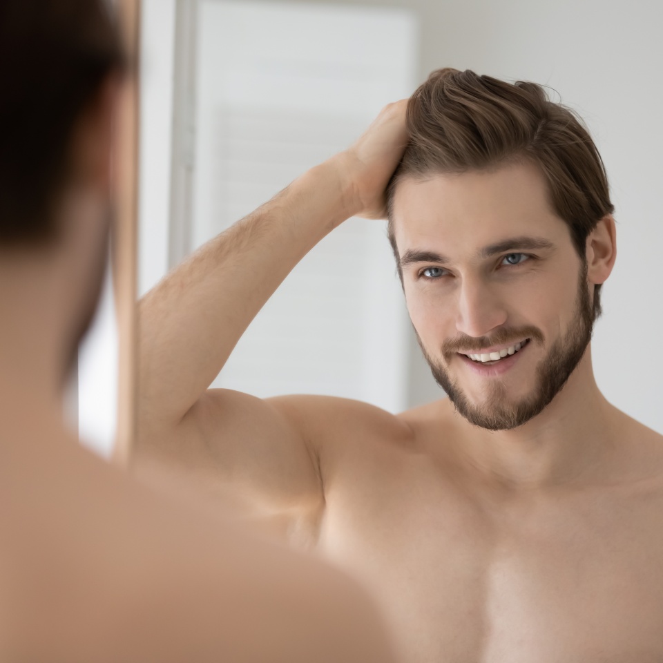 Happy shirtless confident young man looking in mirror combing hair with fingers after morning shower, getting ready for workday or weekend date, feeling satisfied with personal appearance in bathroom.