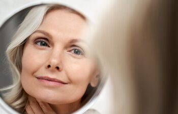 Happy 50s middle aged woman model touching face skin looking in mirror reflection. Smiling mature old lady pampering, healthy moisturized skin care, aging beauty, skincare treatment cosmetics concept.