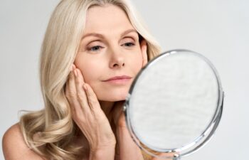 Closeup portrait of gorgeous happy middle age woman looking at mirror touching her skin enjoying treatment for dry skin. Advertising of antiaging beauty skin care products.