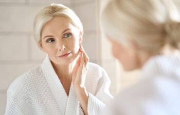 Headshot of gorgeous mid age adult 50 years old blonde woman standing in bathroom after shower touching face, looking at reflection in mirror doing morning beauty routine. Older skin care concept.