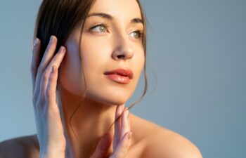 Skin rejuvenation. Aesthetic cosmetology. Facelift treatment. Woman with nude makeup touching flawless radiant face isolated on blue copy space background.