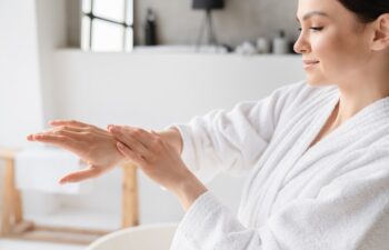 Young caucasian woman in spa bathrobe applying hand cream moisturizer on her hands for softing effect. Recreation, beauty treatment at home