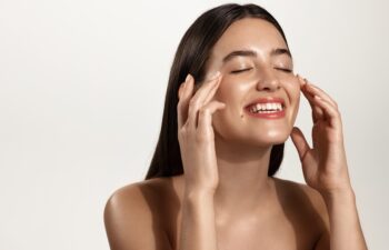 Smiling woman with clean glowing facial skin washing her face, rubbing in cream skincare product for anti-aging, lifting spa effect, standing over white background