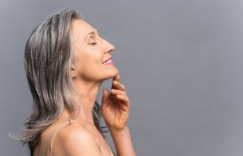 Sensual calm elderly woman stands in profile isolated on gray, smiling with eyes closed, touching chin with hand, gray-haired charming lady enjoying smooth of fresh skin after spa procedure