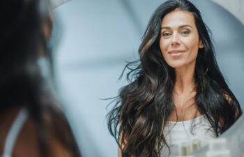 Beautiful Caucasian Middle Aged Woman Looks into Bathroom Mirror, Enjoys Her Looks. Concept for Happiness, Wellbeing, Natural Beauty and Organic Skin Care Products