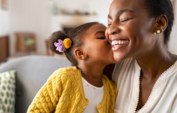 Close up of beautiful daughter kissing mother on cheek at home. African little girl giving kiss to happy mother. Lovely black female child kissing cheerful and proud woman on cheek for mother's day.
