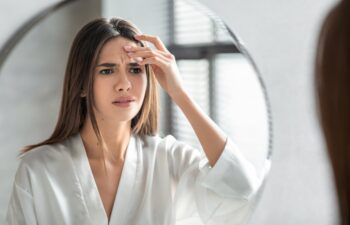 Unhappy Young Woman Looking In Mirror And Touching Wrinkles On Her Face, Attractive Millennial Female Standing In Bathroom And Examining Skin Fine Lines, Selective Focus On Reflection, Closeup