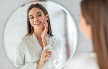 Self-Care Concept. Portrait Of Attractive Young Female Looking At Mirror, Beautiful Woman Wearing White Silk Robe Touching Soft Skin On Face And Smiling, Enjoying Her Reflection, Selective Focus