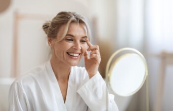 Cheerful beautiful blonde middle aged lady in silky bathrobe using moisturizing eye cream, looking at mirror and smiling, enjoying face care routine, closeup shot. Dark circles, eye wrinkles treatment