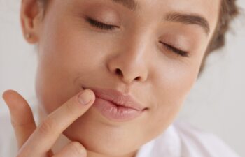 Lips Protection. Beautiful Woman With Happy Face Applying Lip Balm. Portrait of Brunette Girl With Closed Eyes Using Hygienic Balm In Studio. Lips Care Concept
