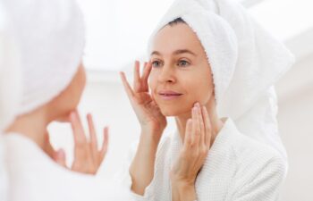 Gorgeous mid age adult 40 years old blonde woman standing in bathroom after shower touching face, looking at reflection in mirror doing morning beauty routine. Older dry skin care concept