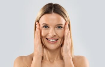 Closeup of happy pretty nude middle aged blonde woman smiling at camera and touching face, posing on grey studio background, pulling face skin, copy space. Plastic surgery, face-lift concept