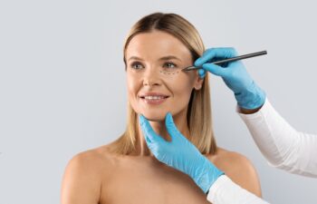 Beautician hands wearing blue medical gloves drawing contours with black pen on woman face before beauty treatment, middle aged blonde lady getting beauty injections, isolated on grey, copy space