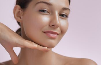 Happy beautiful young adult woman touching her perfect face with healthy shiny skin, hand on chin. Beauty photo of gorgeous girl after morning beauty cosmetic routine finishes daily makeup on pink