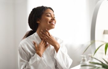 Neck Lift Concept. Smiling African American Woman In Silk Robe Touching Her Soft Smooth Skin While Looking In Mirror At Home, Happy African American Female Enjoying Result Of Beauty Treatments