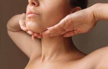 Cropped shot of young caucasian woman touching under the chin with hands massaging her face on dark brown background. Rejuvenation, facelift, facefitness. Exercises from the second chin, pelican neck