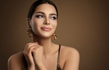 Beauty Model Portrait with plump Lip Makeup looking sideways. Beautiful smiling Woman with smooth Face Skin Make up and Golden Earrings over brown. Dermal Filler