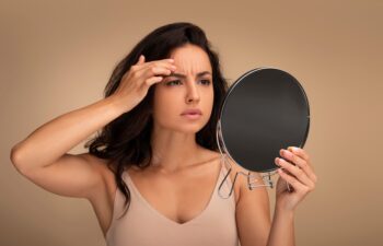 Fine lines and wrinkles, premature skin aging concept. Concerned upset young eastern woman looking in mirror, touching her forehead, checking aging signs on her face, beige background
