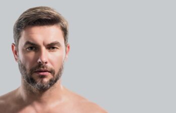 Close up beauty portrait of satisfied man after facial cream. Attractive brunet guy touching skin. Men model face with beard and modern haircut. Banner for header, copy space. Poster for web design.