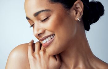 Beauty, skincare and smile with face of woman in studio on gray background for aesthetic or cosmetics. Happy, satisfaction and shoulder with confident model touching skin for dermatology or wellness