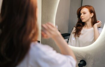 Beautiful young woman in casual clothes, preening, do morning routine, touch her hair standing in front of mirror, smiling admire herself feels satisfied with appearance and complexion.