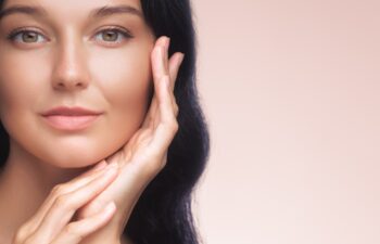 An elegant Asian woman with long black hair gently touches her face, showcasing her flawless skin against a soft pink background. Ideal for beauty, skincare, and cosmetic advertisements, emphasizing