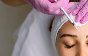 Close up of hands of young cosmetologist injecting botox in female face. She is standing and smiling. The woman is closed her eyes with relaxation