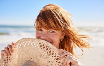Portrait of beautiful woman hiding behind a straw hat at beach and looking at camera with beautiful green eyes. Closeup face of smiling girl with freckles and red hair relaxing at seaside.