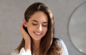 Portrait Of Shy Beautiful Lady With Natural Makeup Posing In Bathroom At Home, Feminine Young Woman With Perfect Smooth Skin Looking Down And Touching Her Hair, Closeup Shot With Copy Space